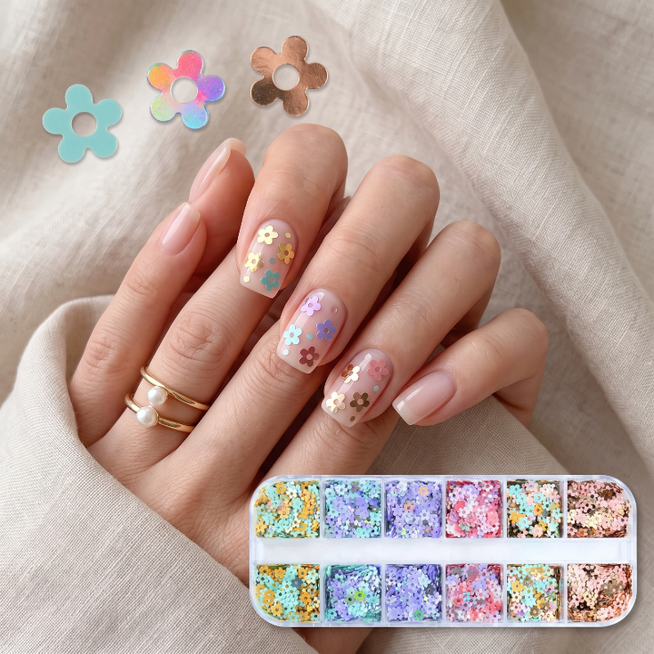 A hand with manicured nails decorated with pastel flower-shaped sequins and glitter, resting on a beige fabric background. A small tray of assorted flower-shaped sequins and glitter is shown at the bottom right, with a few loose flower pieces displayed at the top left.